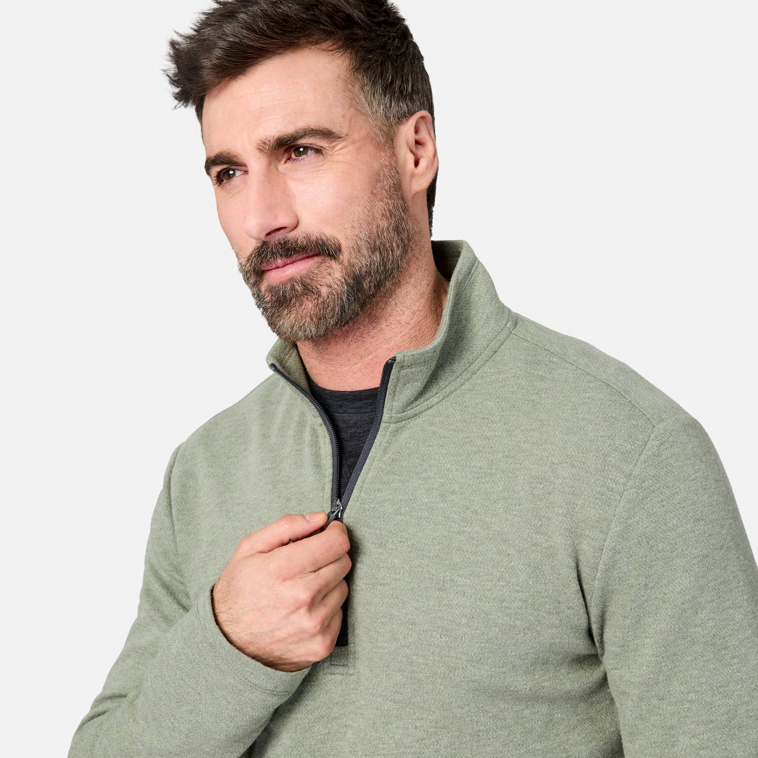 A man with short brown hair and a beard wears the Free Country Men’s Twill Interlock 1/4 Zip Long Sleeve Shirt in a light green color over a dark shirt. He is looking slightly to the side while pulling up the zipper, with a plain, light-colored background behind him.