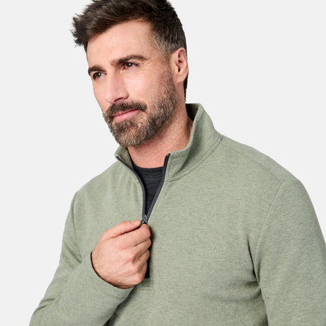 A man with short brown hair and a beard wears the Free Country Men’s Twill Interlock 1/4 Zip Long Sleeve Shirt in a light green color over a dark shirt. He is looking slightly to the side while pulling up the zipper, with a plain, light-colored background behind him.