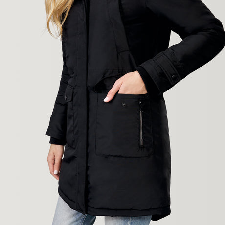 A person wearing the Clearance Women's Arctic Anorak Jacket—a long black eco-friendly coat with a front zipper and flap pockets—has their hand in one pocket, paired with light blue jeans and shown from the shoulders down.