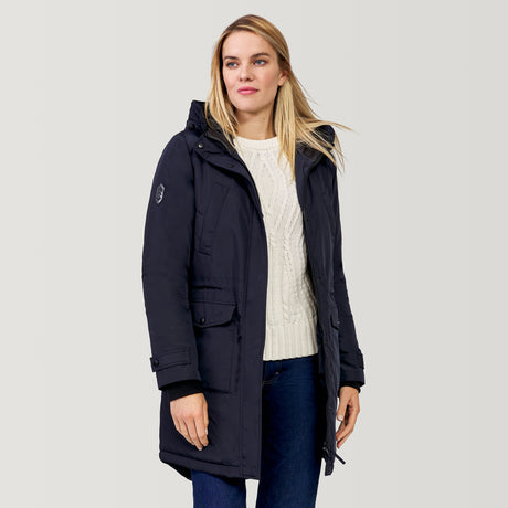 A woman wears the Clearance Women's Arctic Anorak Jacket, a dark navy wind-resistant coat with REPREVE fill, over a white cable-knit sweater and blue jeans, standing against a plain light gray background and gently smiling.