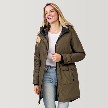 A smiling woman with long blonde hair models the Clearance Women's Arctic Anorak Jacket—a green eco-friendly parka with REPREVE fill—over a white shirt and light blue jeans against a plain light background.