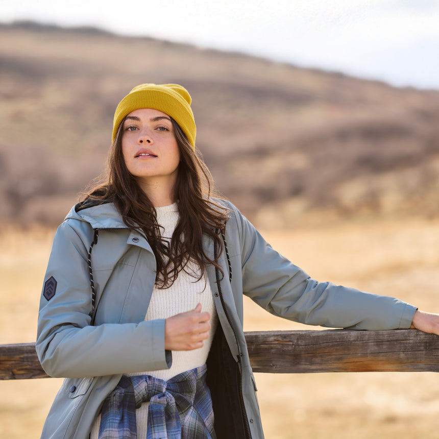 Woman wearing a yellow beanie and green coat standing by a wooden fence in a field.