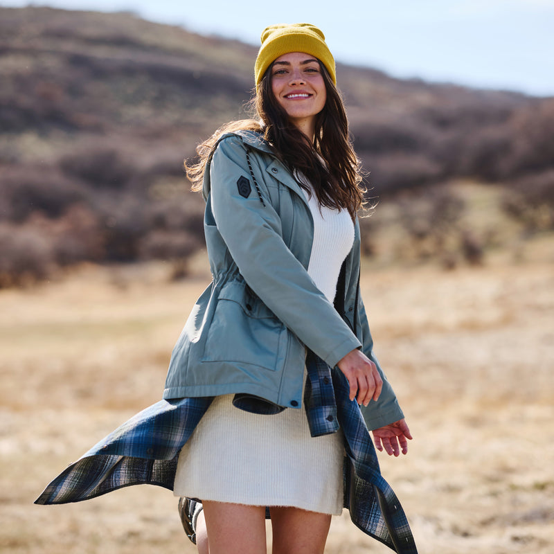 Woman in a green jacket and yellow beanie standing in a desert-like landscape