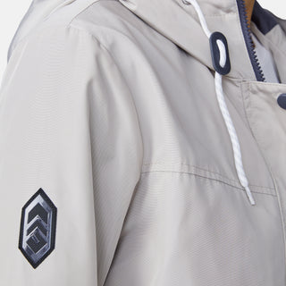 Close-up of the Free Country Women's Rain Away Anorak Rain Jacket in light beige, featuring a white drawstring hood, black geometric sleeve patch, and partially visible zipper and snap button details.