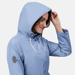 A woman smiles, adjusting the hood of her light blue Free Country Women's Rain Away Anorak Rain Jacket. The water-resistant jacket features white drawstrings and a black logo patch on the sleeve against a plain white background.