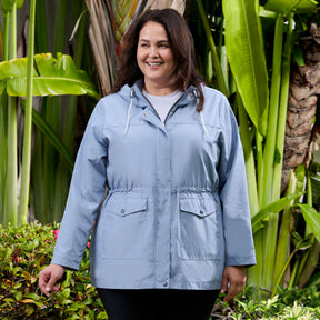 A woman wearing the Free Country Women's Plus Size Rain Away Anorak Rain Jacket in light blue smiles outdoors, surrounded by lush green plants and tropical foliage.