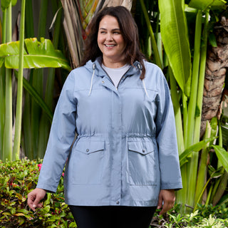A woman wearing the Free Country Women's Plus Size Rain Away Anorak Rain Jacket in light blue smiles outdoors, surrounded by lush green plants and tropical foliage.