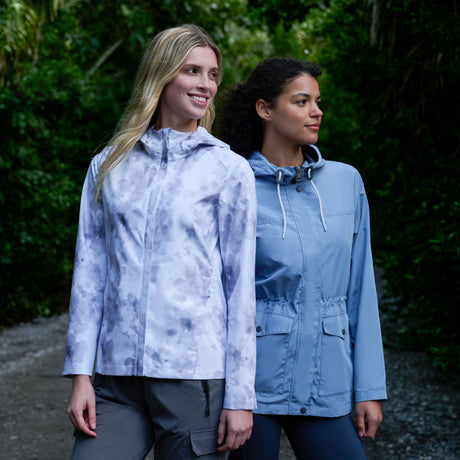 Two women stand outdoors on a forest path. One wears gray pants and a patterned white jacket; the other wears the Free Country Women's X2O Packable Rain Jacket—stylish, waterproof, and offering UPF 50+ protection. Both are smiling and looking off-camera.