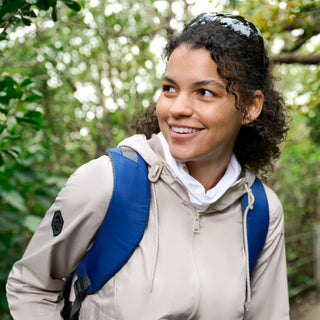 A woman with curly hair smiles in a lush green forest, wearing the Free Country Women's Misty Rain Away Anorak Rain Jacket and a blue backpack. Sunglasses rest on her head.