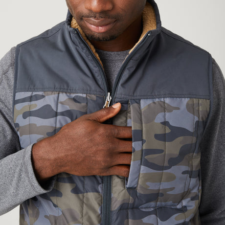 A man wears the Men's FreeCycle® Alpine Reversible Vest by Clearance—a blue camo sherpa vest with a fleece collar—over a gray long-sleeve shirt, touching its zippered chest pocket. Only his torso and lower face are shown.