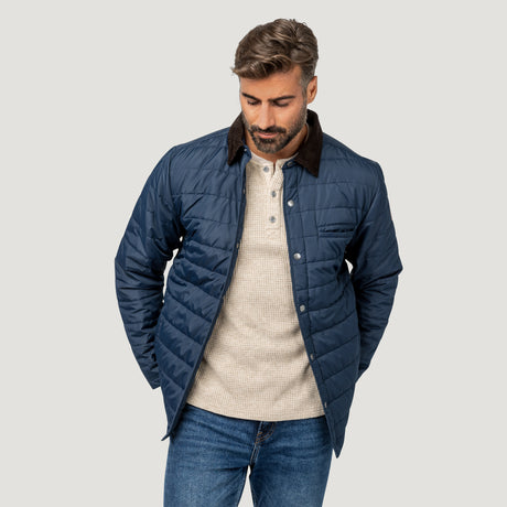[Model is wearing size M.] A man wearing a navy blue quilted jacket with a dark brown corduroy collar, layered over a beige knit polo shirt and blue jeans, set against a solid light gray background.