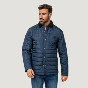[Model is wearing size M.] A man wearing a navy blue quilted jacket with a dark brown corduroy collar, paired with blue jeans, photographed against a solid light gray background.