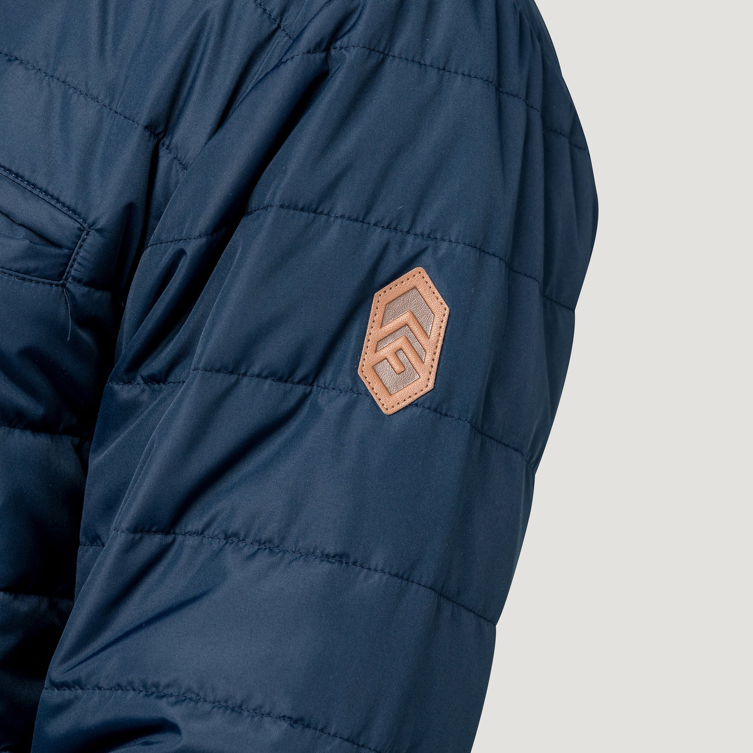 [Model is wearing size M.] Close-up of a navy blue quilted puffer jacket sleeve featuring a tan hexagonal patch with a stylized 'F' logo on the upper arm, set against a light gray background.