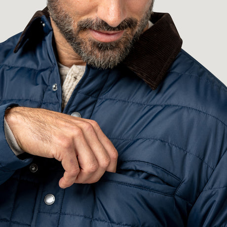 [Model is wearing size M.] Close-up of a man wearing a navy blue quilted jacket with a dark brown corduroy collar, showing his hand in the pocket and part of his beard and shirt.