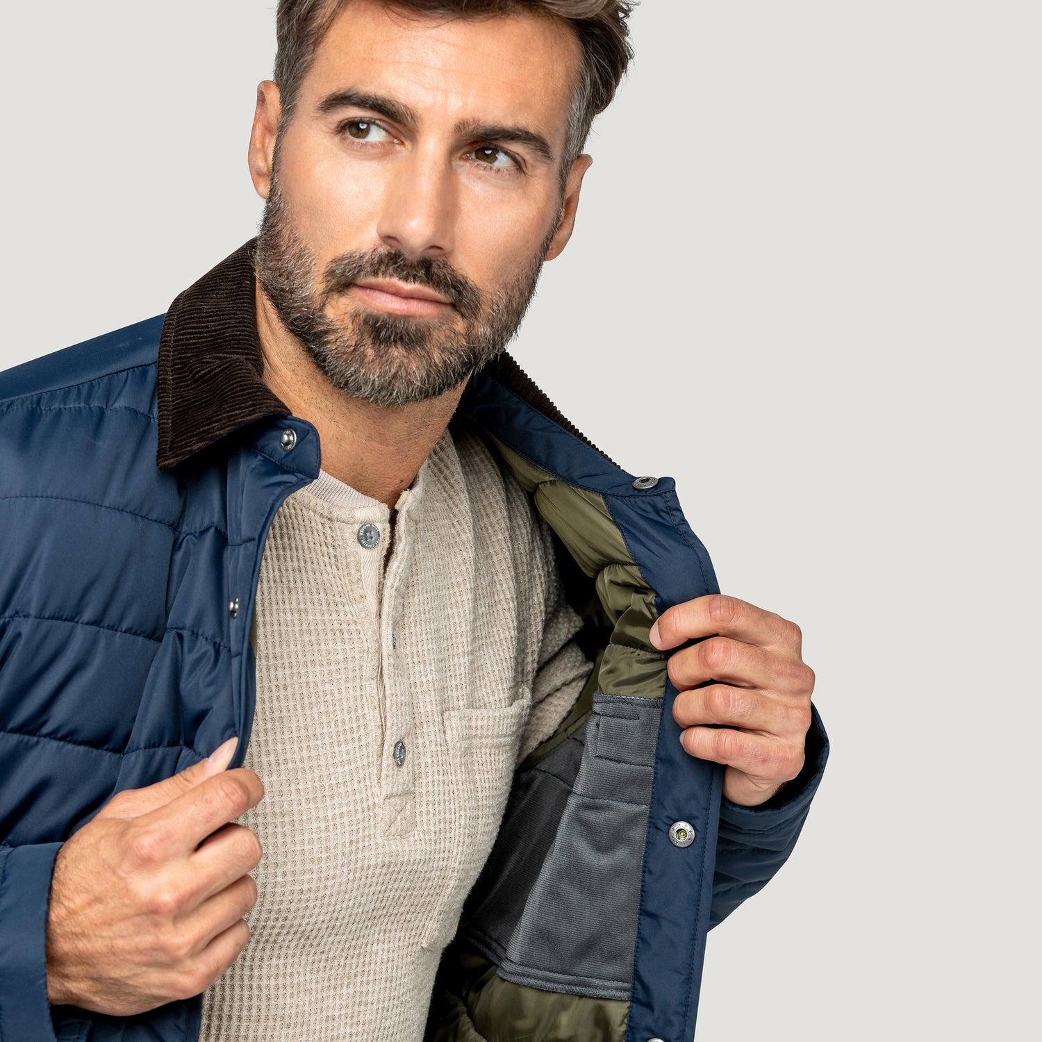 [Model is wearing size M.] A close-up of a man wearing a navy blue puffer jacket with a brown corduroy collar, unzipped to show a gray and olive green lining, over a beige henley shirt, against a light gray background.