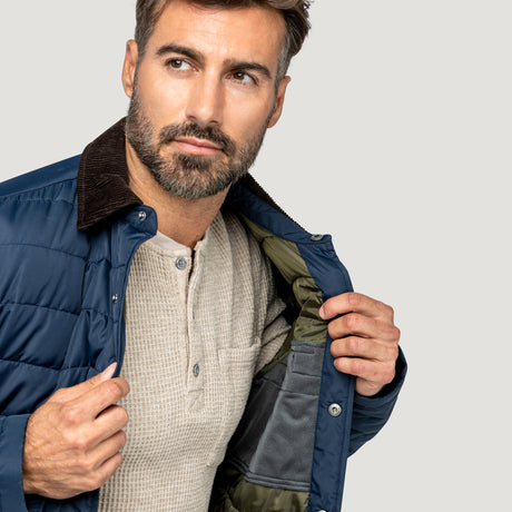 [Model is wearing size M.] A close-up of a man wearing a navy blue puffer jacket with a brown corduroy collar, unzipped to show a gray and olive green lining, over a beige henley shirt, against a light gray background.