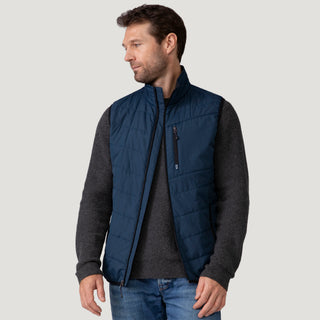[Vince is 6’1” wearing a size Medium.] Man wearing the Free Country Men's FreeCycle® Stimson Puffer Vest in navy, featuring quilted design, REPREVE® poly fill, and a vertical zip, layered over a burgundy long-sleeve shirt with dark jeans.