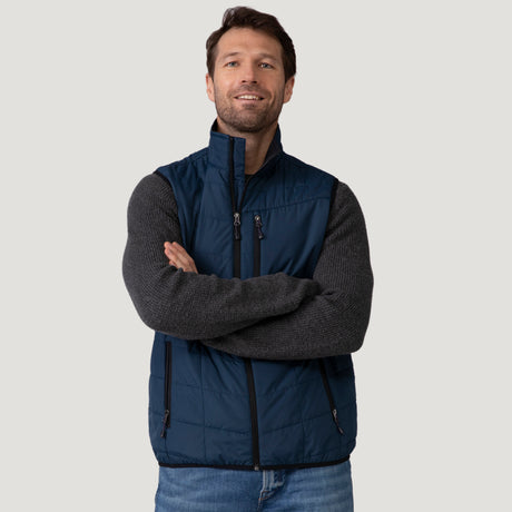 [Vince is 6’1” wearing a size Medium.] Man wearing the Free Country Men's FreeCycle® Stimson Puffer Vest in navy, featuring quilted design, REPREVE® poly fill, and a vertical zip, layered over a burgundy long-sleeve shirt with dark jeans.