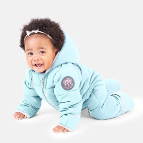 A smiling baby in the Clearance Kids' Out of Bound Puffer Snow Suit with a sherpa-lined hood and a white headband crawls on a white surface.