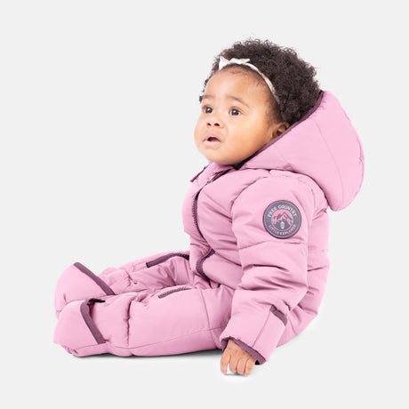 A young child with curly hair sits on the floor in a light pink Kids' Out of Bound Puffer Snow Suit by Clearance, featuring matching mittens and booties, and cozy recycled polyfill, looking slightly up and to the side.