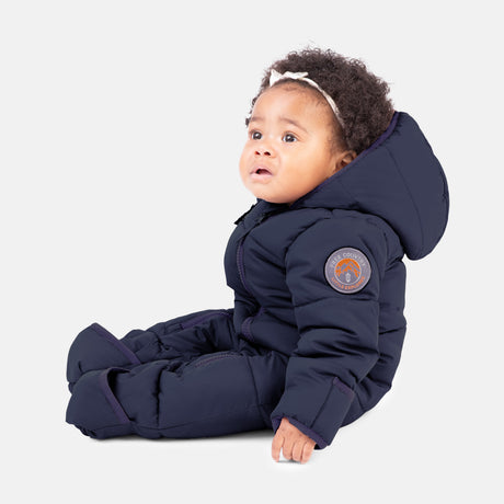 A baby with curly hair in a Clearance Kids' Out of Bound Puffer Snow Suit, featuring sherpa lining and double zippers, sits on the floor in front of a plain white background, wearing a light headband.