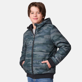A boy smiles at the camera in the Clearance Boys' Poofie Reversible Puffer Jacket with a camo pattern and hood. His hands are in its pockets, paired with blue jeans, against a plain white background. The jacket is made from recycled polyester.