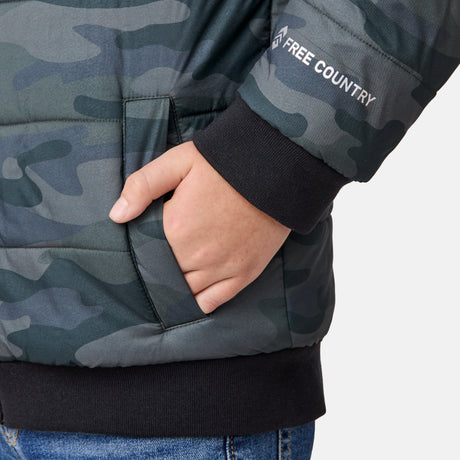 A person wears a Boys' Poofie Reversible Puffer Jacket by Clearance in green and gray camo, blue jeans, and has their right hand in the jacket pocket against a light background.