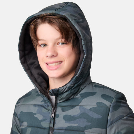 A smiling teenage boy wears the Boys' Poofie Reversible Puffer Jacket by Clearance, featuring a camo-patterned hood, standing against a plain light background.