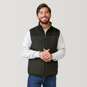 A man with brown hair and a beard smiles while wearing a light gray sweater, blue jeans, and the Clearance Men's FreeCycle® Alpine Reversible Vest with recycled polyfill, standing against a plain light gray background.