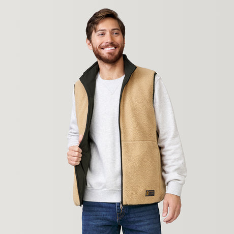 A smiling man with brown hair and a beard wears the Clearance Men's FreeCycle® Alpine Reversible Vest over a light gray sweatshirt and blue jeans, standing against a plain light background.