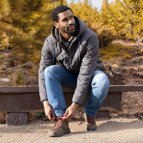 Man sitting on a wooden bench in a forested area, tying his shoe.