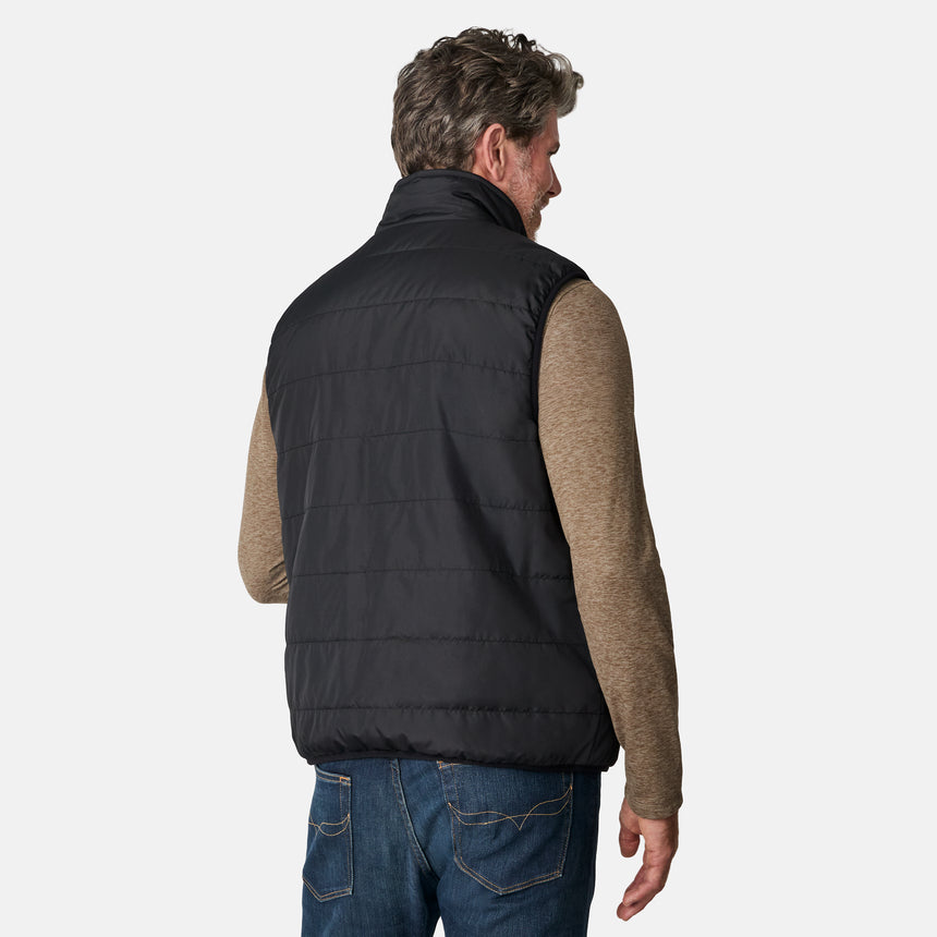 [Kolby is 6'2" wearing size M.] Free Country men's reversible black quilted vest with zip-front closure and side pockets—worn over a beige long-sleeved shirt and blue jeans for a layered, transitional-weather look.