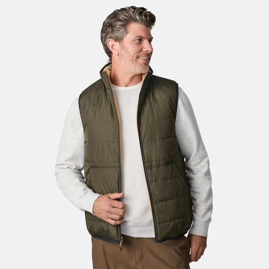 [Kolby is 6'2" wearing size M.] Men’s Free Country green quilted vest with zip-front closure and stand collar—reversible to tan sherpa fleece for versatile warmth and style. Shown layered over a light gray crewneck shirt with brown pants, styled for rugged, outdoor-ready appeal.