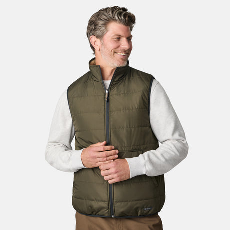 [Kolby is 6'2" wearing size M.] Men’s Free Country green quilted vest with zip-front closure and stand collar—reversible to tan sherpa fleece for versatile warmth and style. Shown layered over a light gray crewneck shirt with brown pants, styled for rugged, outdoor-ready appeal.