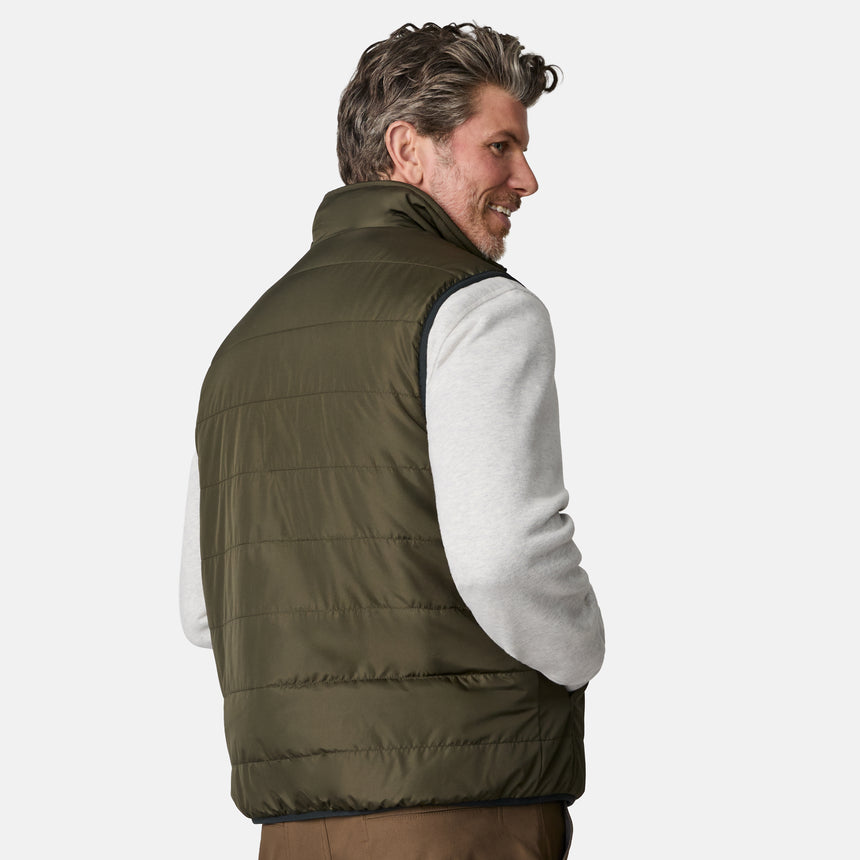 [Kolby is 6'2" wearing size M.] Men’s Free Country green quilted vest with zip-front closure and stand collar—reversible to tan sherpa fleece for versatile warmth and style. Shown layered over a light gray crewneck shirt with brown pants, styled for rugged, outdoor-ready appeal.