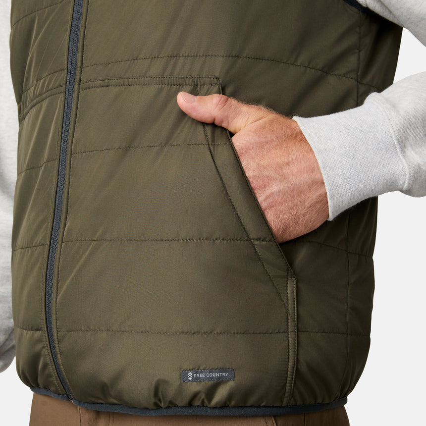 [Kolby is 6'2" wearing size M.] Men’s Free Country green quilted vest with zip-front closure and stand collar—reversible to tan sherpa fleece for versatile warmth and style. Shown layered over a light gray crewneck shirt with brown pants, styled for rugged, outdoor-ready appeal.