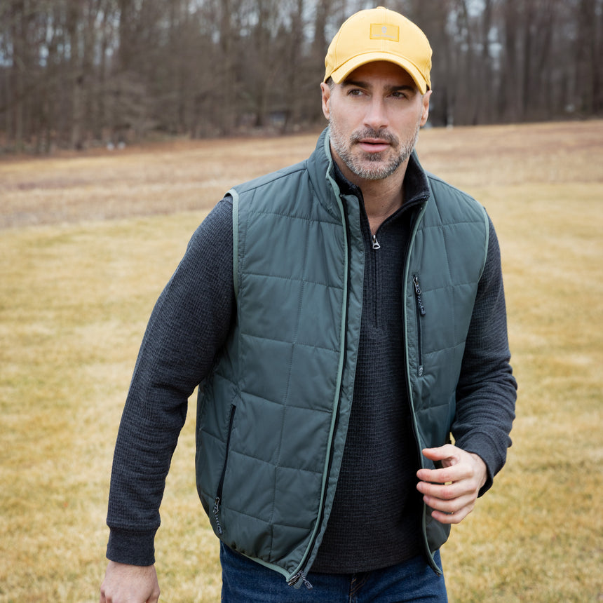 Man wearing a green puffer vest and yellow cap standing in a field.