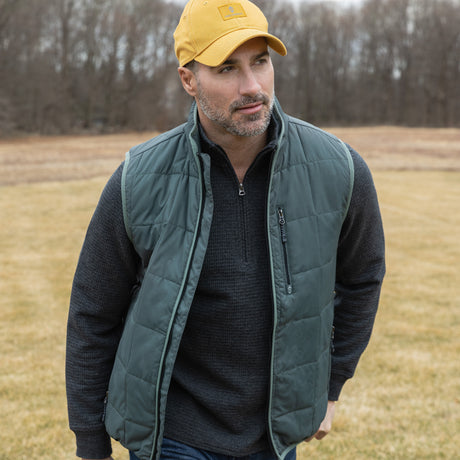 Man wearing a green puffer vest and yellow cap standing in a field.