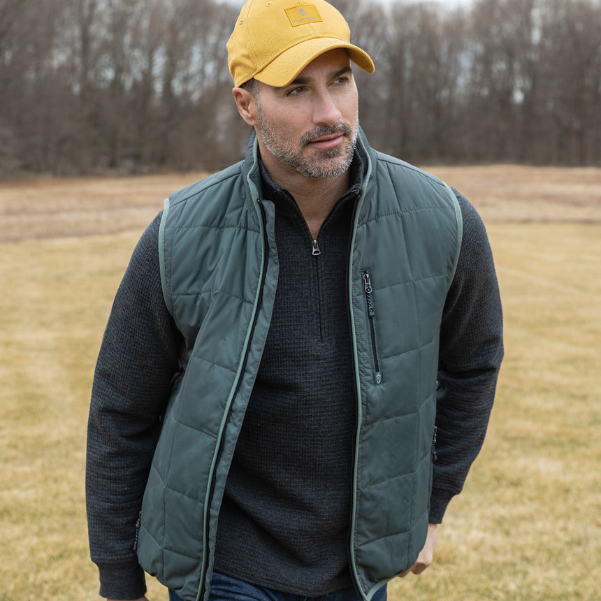 Man wearing a green puffer vest and yellow cap standing in a field.