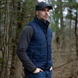 Man wearing a blue puffer vest and black cap standing in a forest.