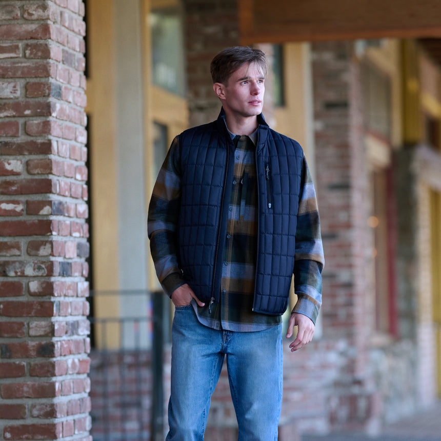 A man wearing a navy quilted zip-front puffer vest over a brown and blue plaid flannel shirt, paired with blue jeans, standing in a brick corridor.