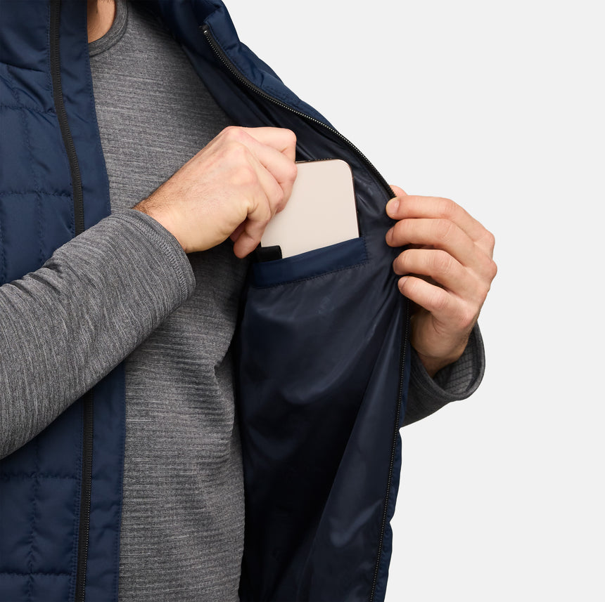 Person wearing navy Sperrin full zip puffer vest over a grey knit, holding a smartphone at chest level.