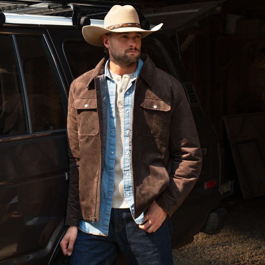 A man wearing a brown Men’s Faux Suede Full Zip Shirt Jacket over a denim shirt and a cowboy hat stands next to a dark SUV.