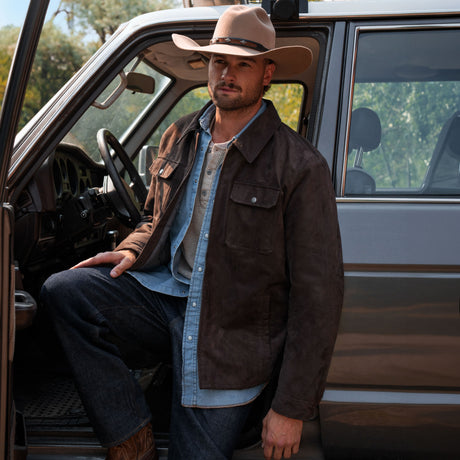 A man wearing a Free Country Men’s Faux Suede Full Zip Shirt Jacket over a light blue shirt and a cowboy hat stands next to an open car door.
