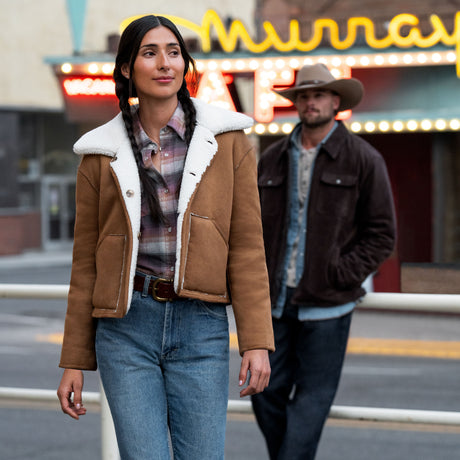 A woman wearing a tan Free Country Women’s Faux-Suede Shearling Reversible jacket stands in front of a man in a cowboy hat and dark jacket outside a cafe.