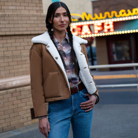 A woman wearing a tan Free Country Women’s Faux-Suede Shearling Reversible Jacket over a plaid shirt and blue jeans.
