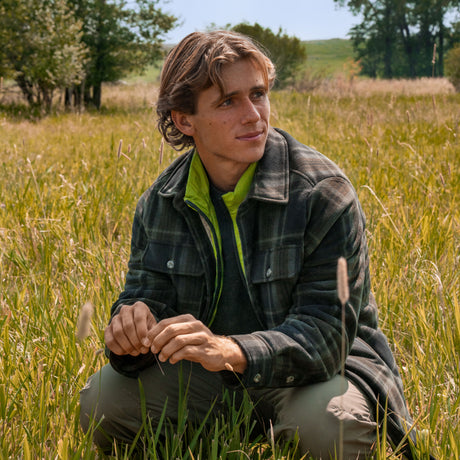 A man wearing a green plaid Men’s Mountain Peak Chill Out Fleece Shirt Jacket by Free Country crouches in a grassy field.