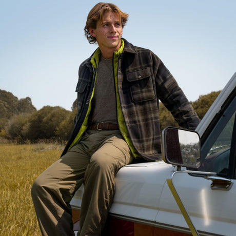 A man wearing a Free Country Men’s Mountain Peak Chill Out Fleece Shirt Jacket over a lime green vest sits on the hood of a truck in a field.
