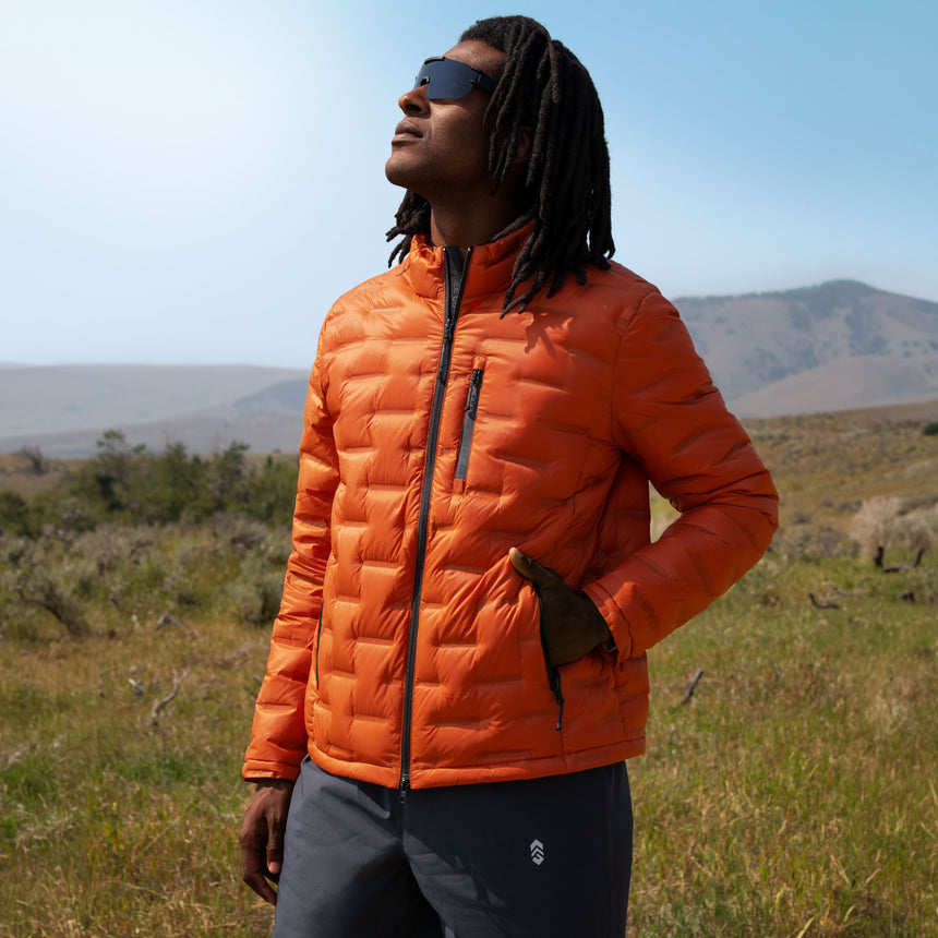 A person wearing an orange, lightweight, packable Men’s Paramount Down Packable Puffer Jacket and dark sunglasses stands in a grassy field.