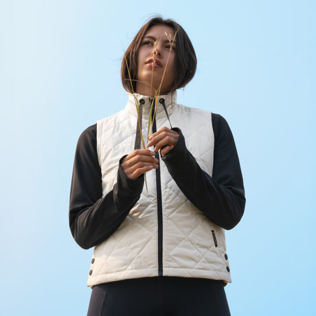 A woman wearing a white Free Country Women’s Cirrus Diamond Quilted Vest over a black long-sleeved shirt, holding a sprig of grass against a light blue sky.